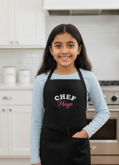 Young girl wearing a black apron with 'CHEF Maya' embroidered text in a kitchen setting