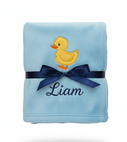 Folded blue blanket with a yellow duck and navy ribbon, and personalized embroidery with 'Liam'.