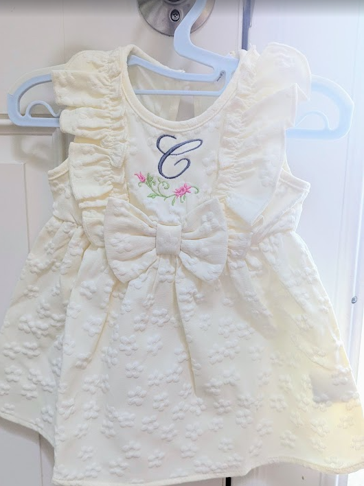White baby dress with a bow and embroidered monogram letter 'C' on a hanger.