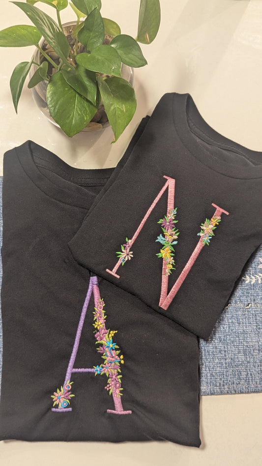 Black t-shirt with colorful embroidered letters on a textured surface with a plant in the background