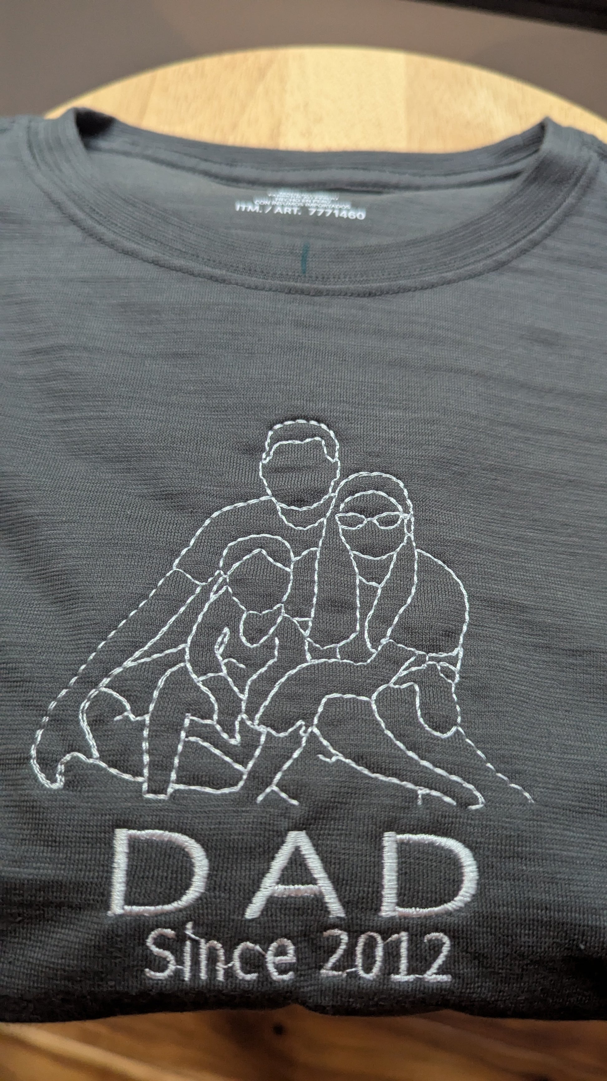 Gray t-shirt with a graphic design and 'DAD Since 2012' and an outline picture embroidered  on a wooden surface