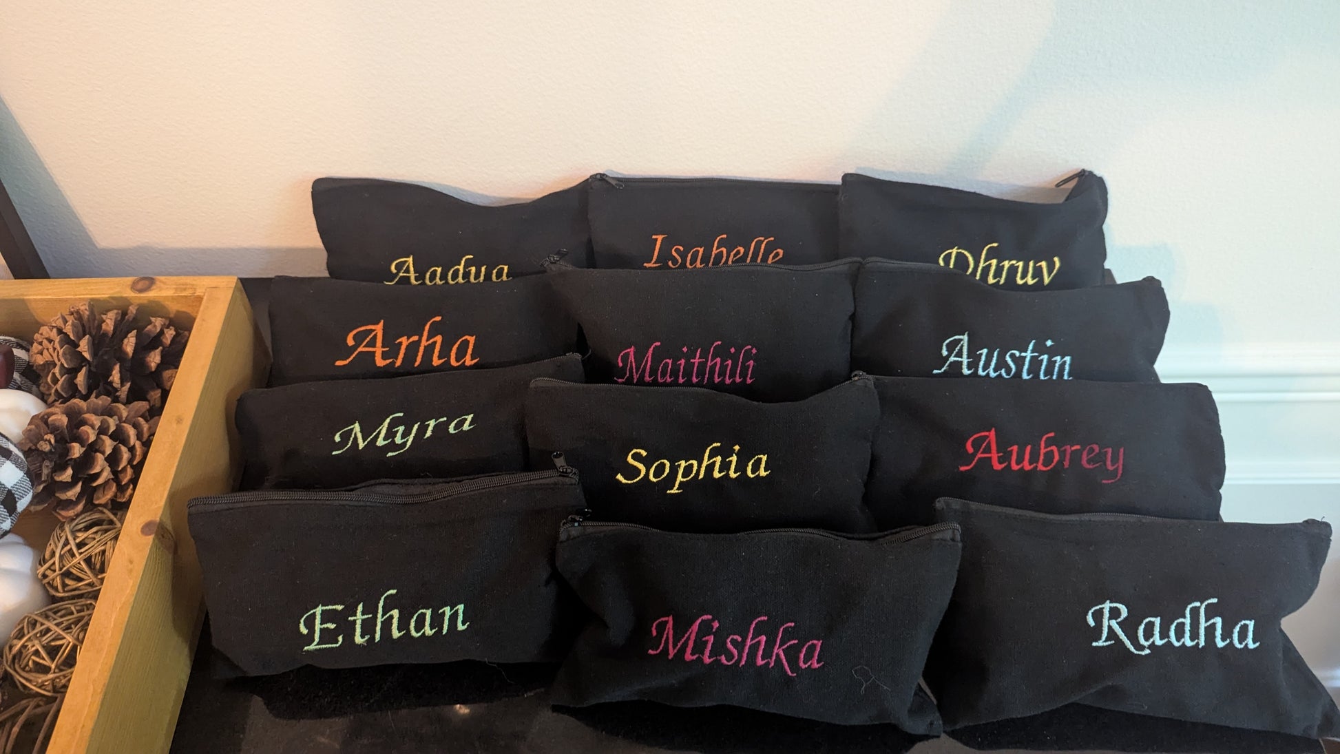 Black pouches with embroidered names in various colors on a wooden shelf.