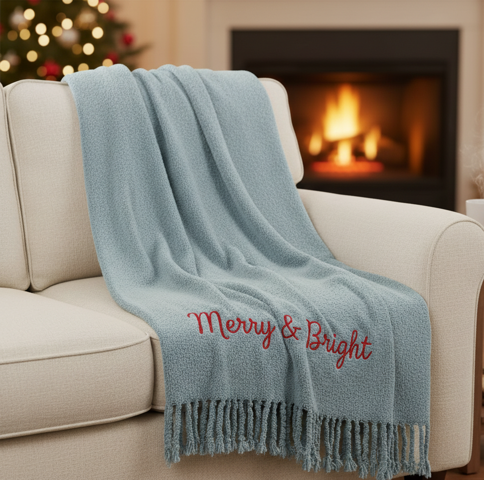 Light blue blanket with 'Merry & Bright' text embroidered draped over a beige sofa in front of a fireplace.