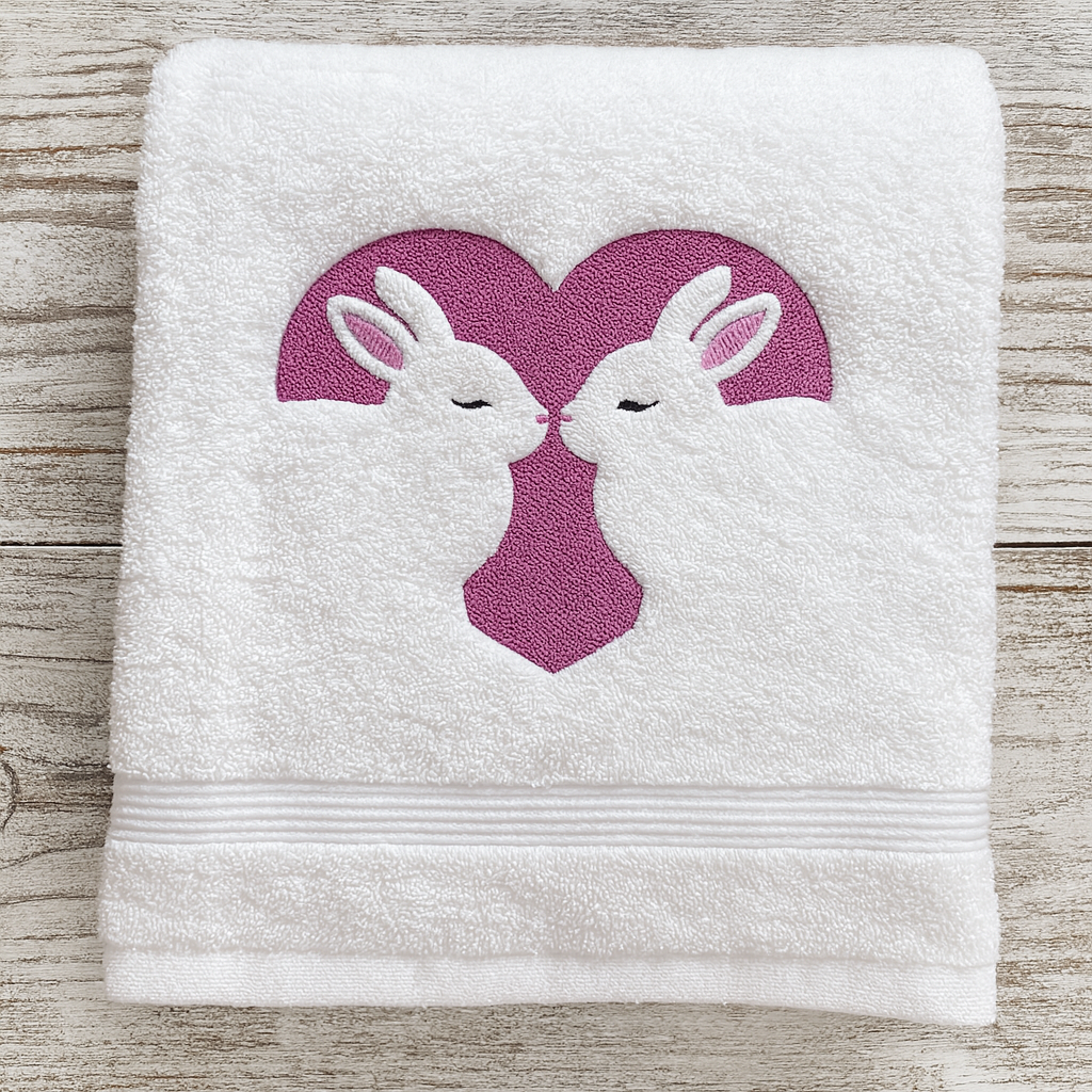 White towel with embroidered design of two rabbits in a heart on a wooden surface